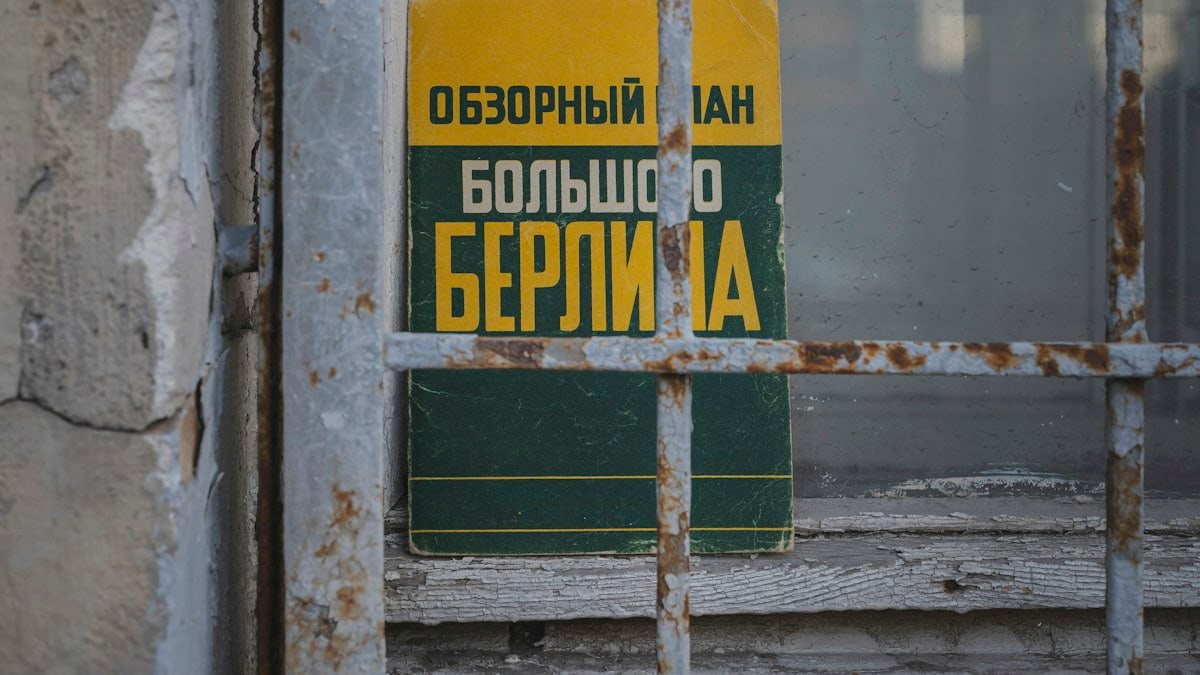 A weathered guidebook to Greater Berlin sits behind a rusty window grille on a cracked wooden sill — its yellow-and-green cover and bold Cyrillic type peeking out from a quiet, forgotten storefront. The composition pairs vintage travel optimism with urban decay: peeling paint, textured metal, and soft, muted light create a nostalgic, cinematic study of place, memory, and small found objects.